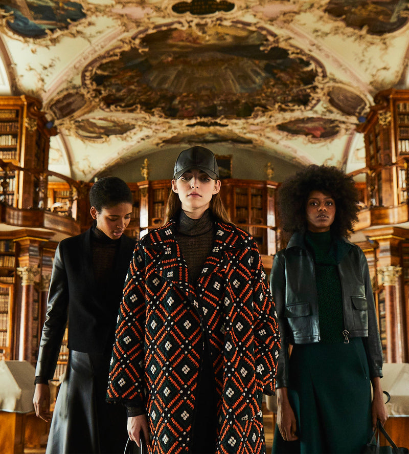 Drei Models in der Stiftsbibliothek in St. Gallen, ein Model trägt einen mehrfarbigen Mantel aus feinster Stickerei. Im Hintergrund kann die reich dekorierte Decke bestaunt werden.