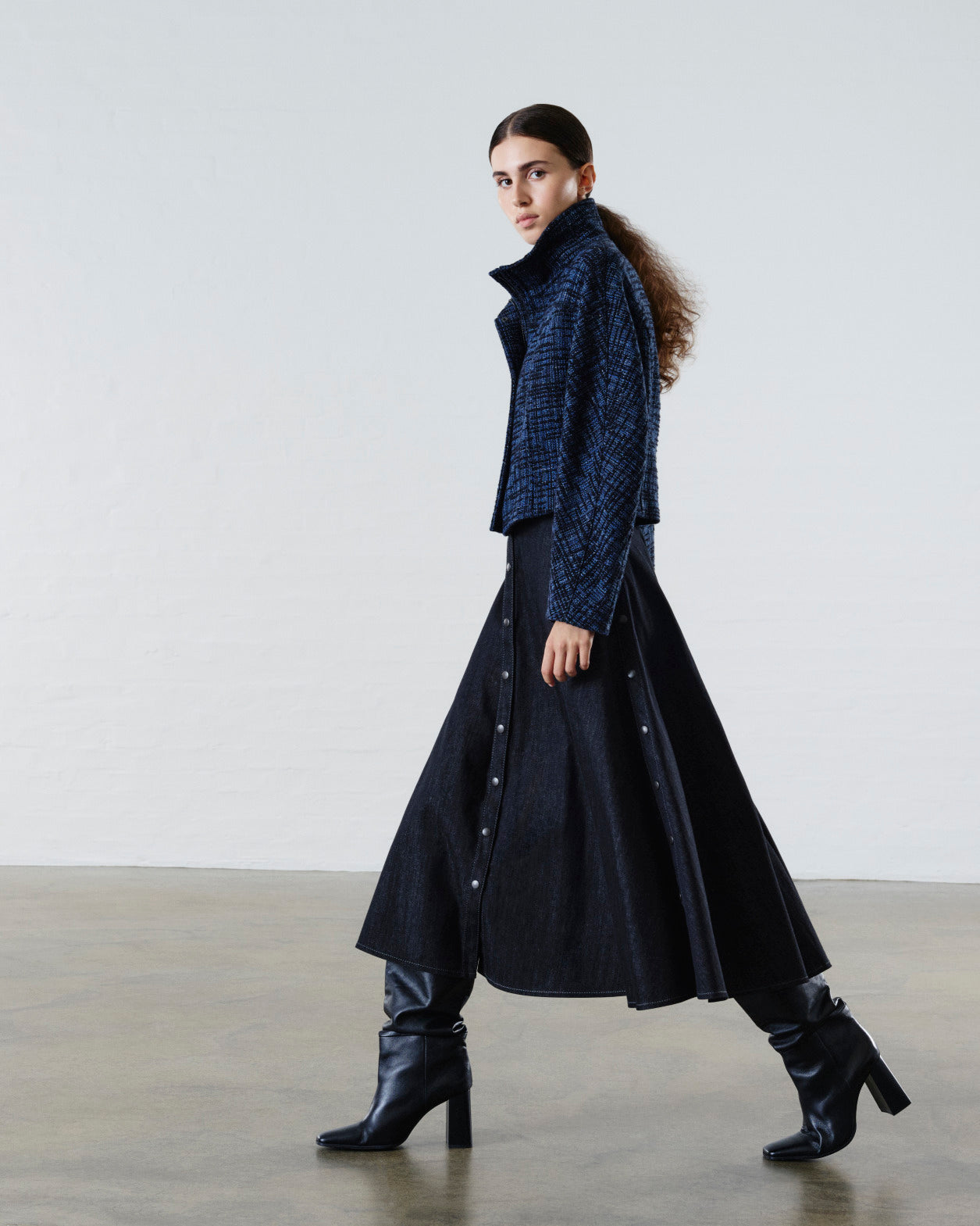 Luxuriöser Damenlook mit strukturierter blauer Cropped-Jacke, schwarzem ausgestelltem Midirock und schwarzen Stiefeletten mit Absatz, präsentiert an einem weiblichen Model in einem minimalistischen Studio.