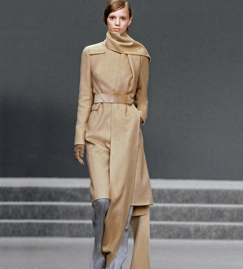 Model auf dem Laufsteg trägt einen langen beigen Mantel und einen langen Schal in derselben Farbe.