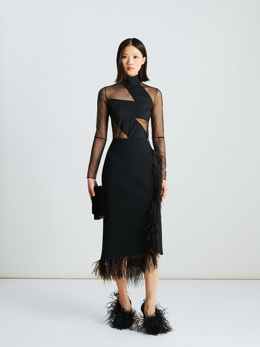Akris Fall/Winter look featuring a sheer fitted mock-neck top with zigzag detail in black, paired with an asymmetrical wool midi skirt with feather trim — a sleek ensemble blending graphic precision and fluid movement.