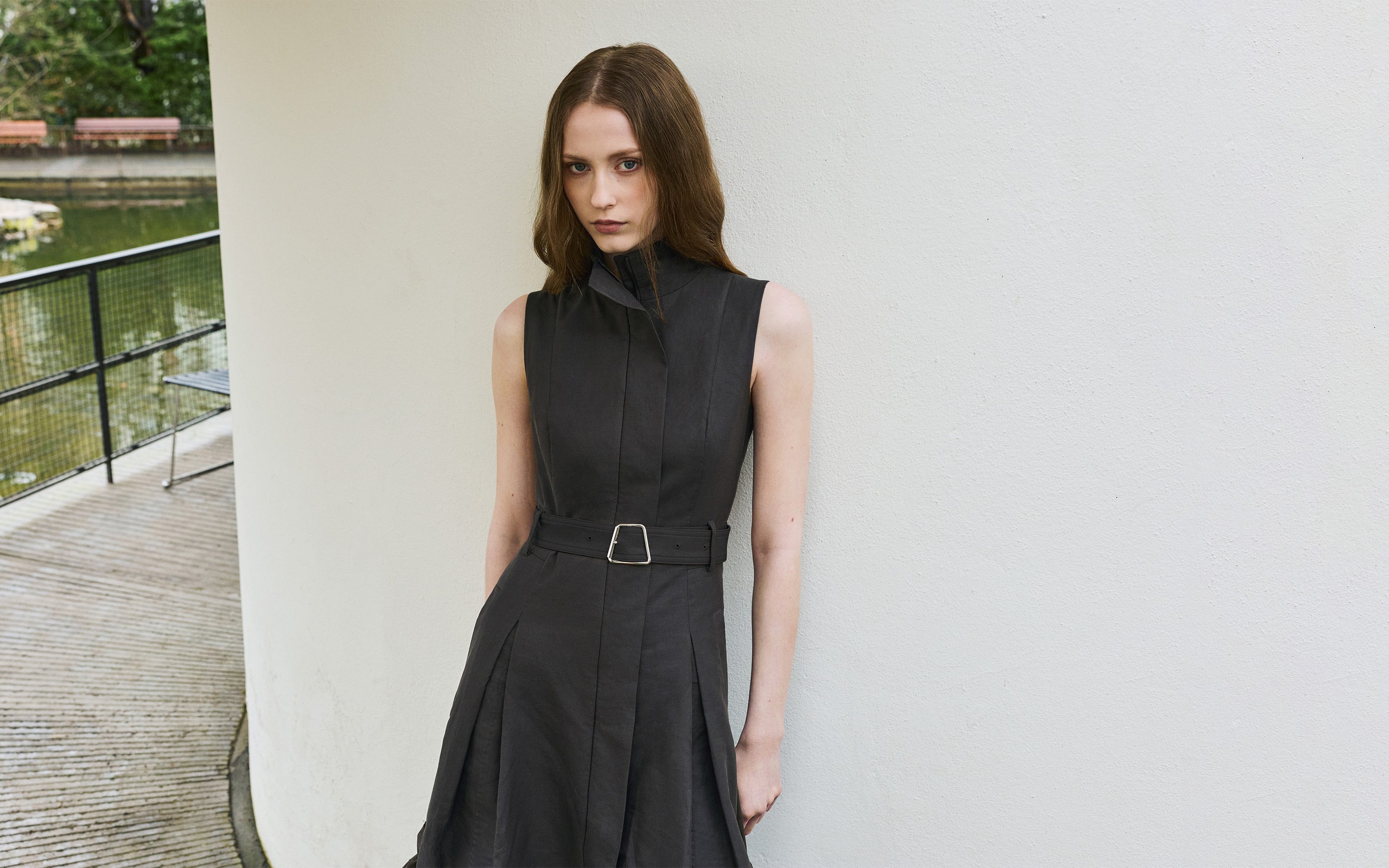 Akris Frühjahr 2026 Kollektion: Modell in einem ärmellosen, strukturierten Kleid in Dunkelgrau mit Stehkragen und Gürtel, an einer weißen Wand lehnend.