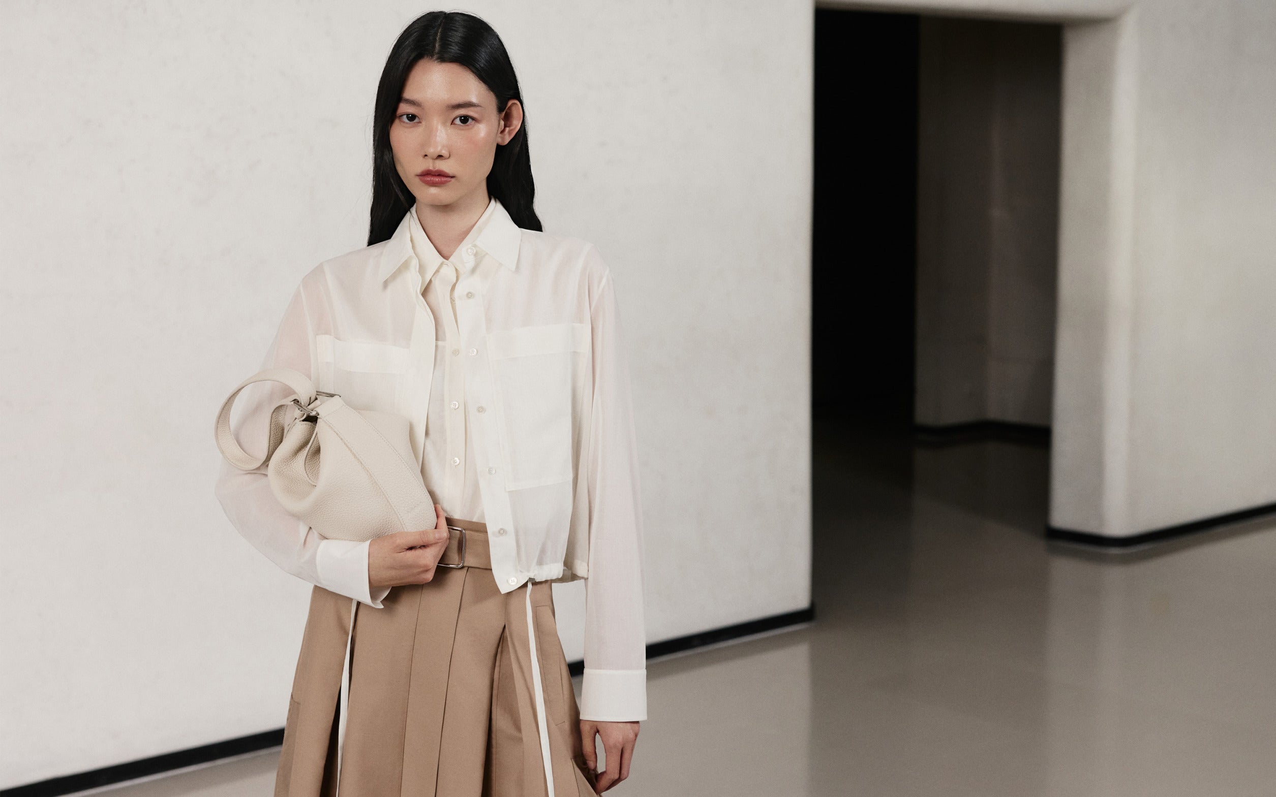 Model trägt eine weiße Seidenbluse, einen beigen Rock und eine elfenbeinfarbene Ledertasche aus der Akris Pre-Fall 2026 Kollektion.