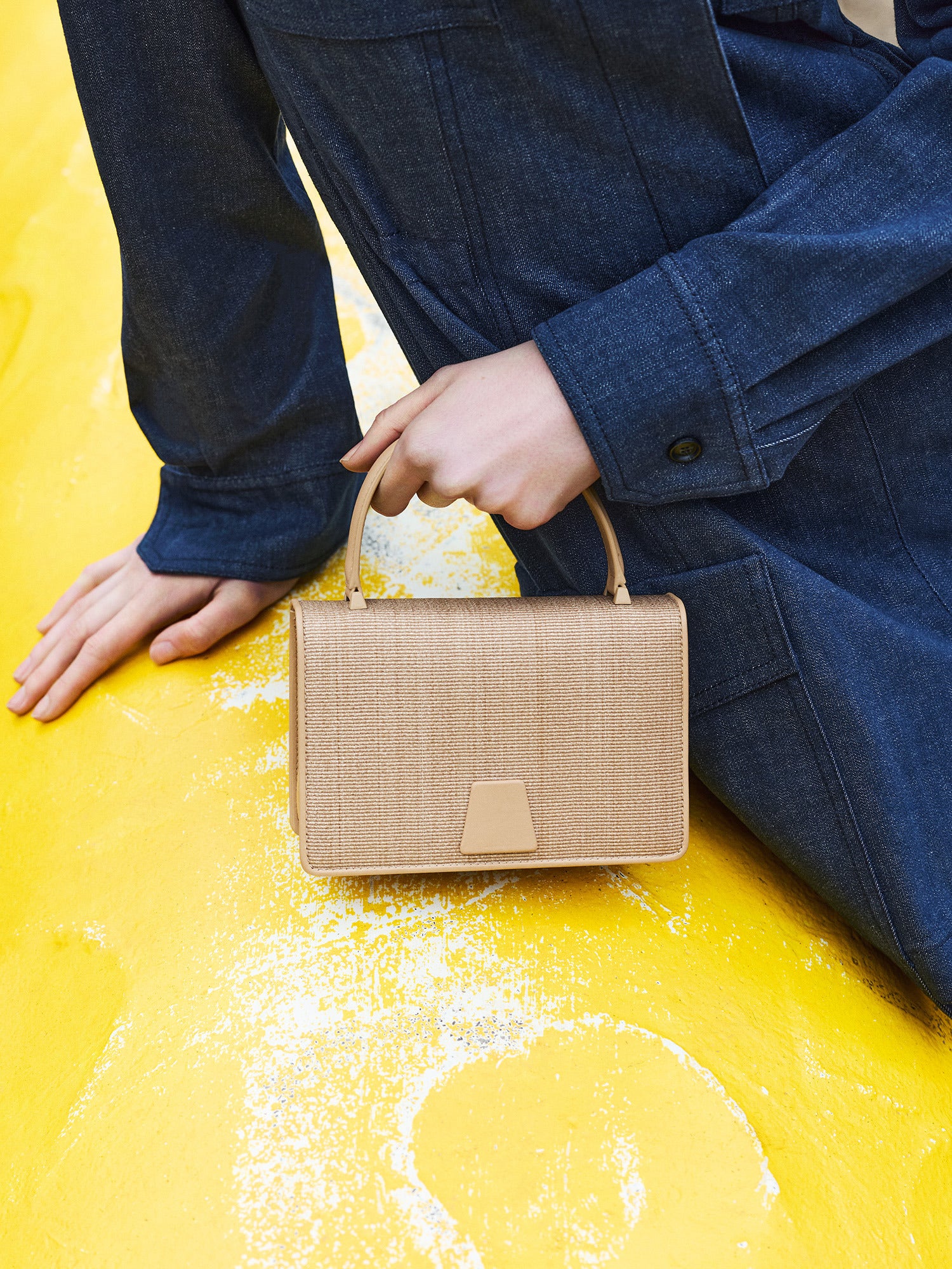 Ein Modell trägt eine dunkelblaue Denimjacke von Akris und sitzt auf einer gelben Fläche, während es eine Akris Ai Handtasche hält. Die Tasche ist aus natürlichem Strohgeflecht gefertigt, hat einen hellbraunen Ledergriff und einen trapezförmigen Verschluss aus Leder auf der Vorderseite.