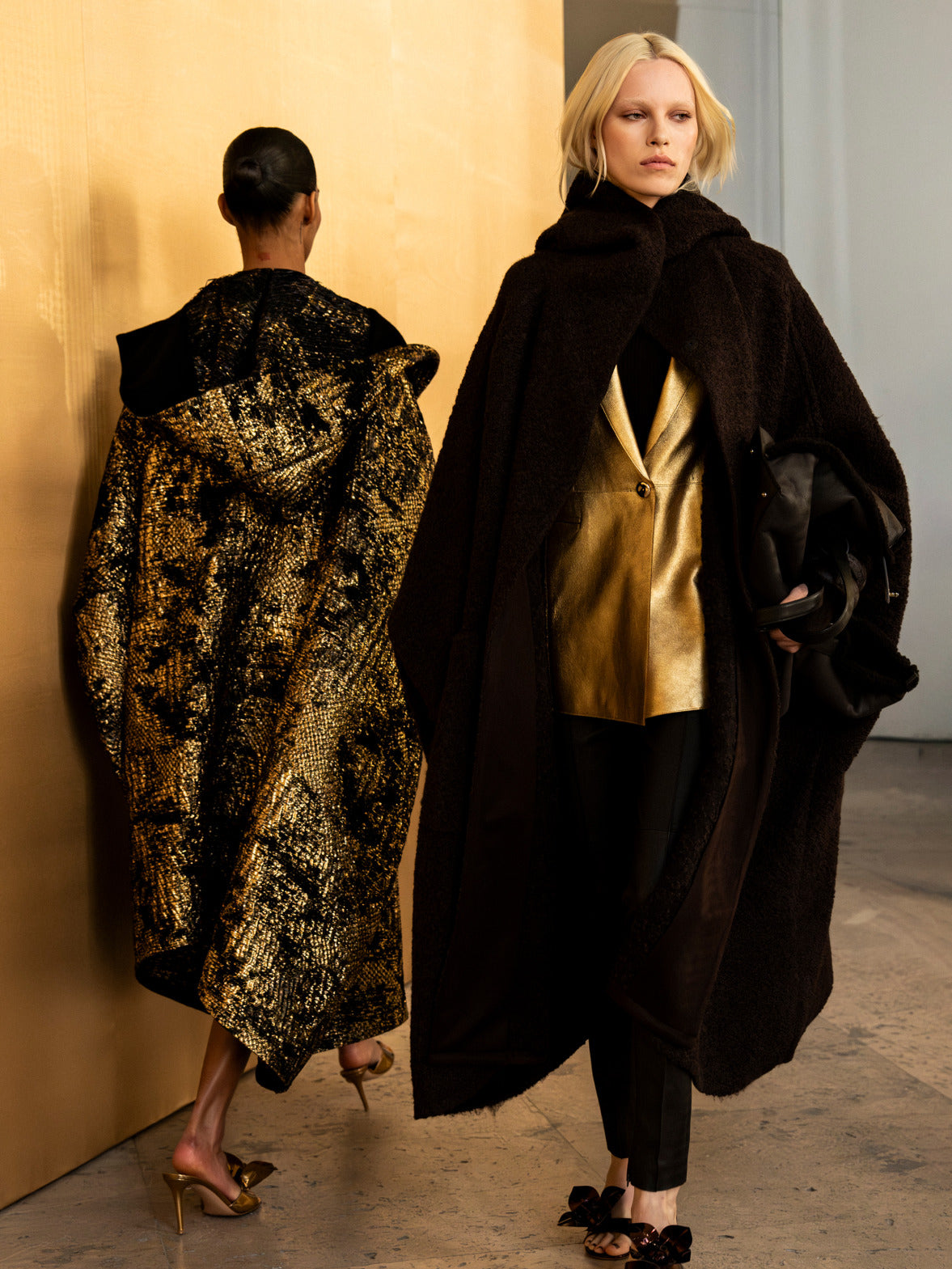 Zwei Models präsentieren luxuriöse Texturen, darunter ein voluminöser dunkler Strukturmantel über einem goldenen Metallic-Blazer und ein schimmerndes Cape mit Goldmuster.