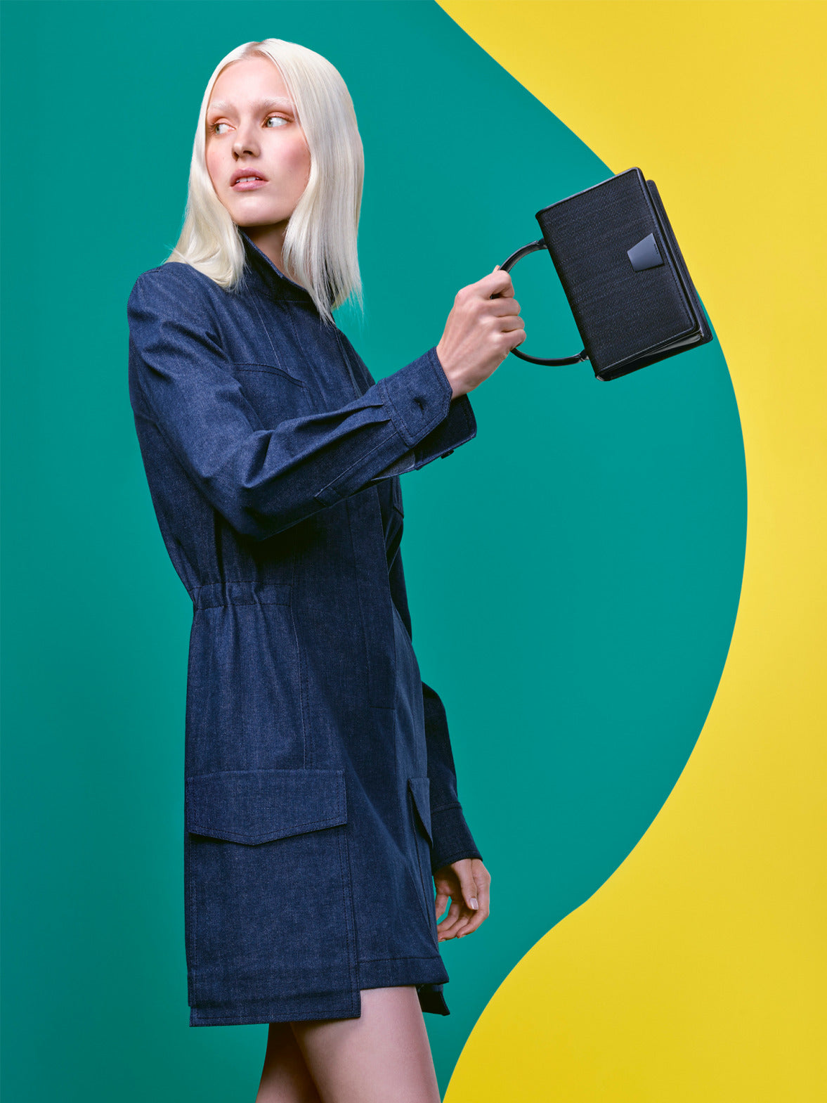 Ein Model mit platinblonden Haaren in einem dunklen Denim-Mantelkleid, das eine strukturierte schwarze Handtasche hält.
Der Hintergrund besteht aus einem minimalistischen Color-Blocking-Design in Grün und Gelb.