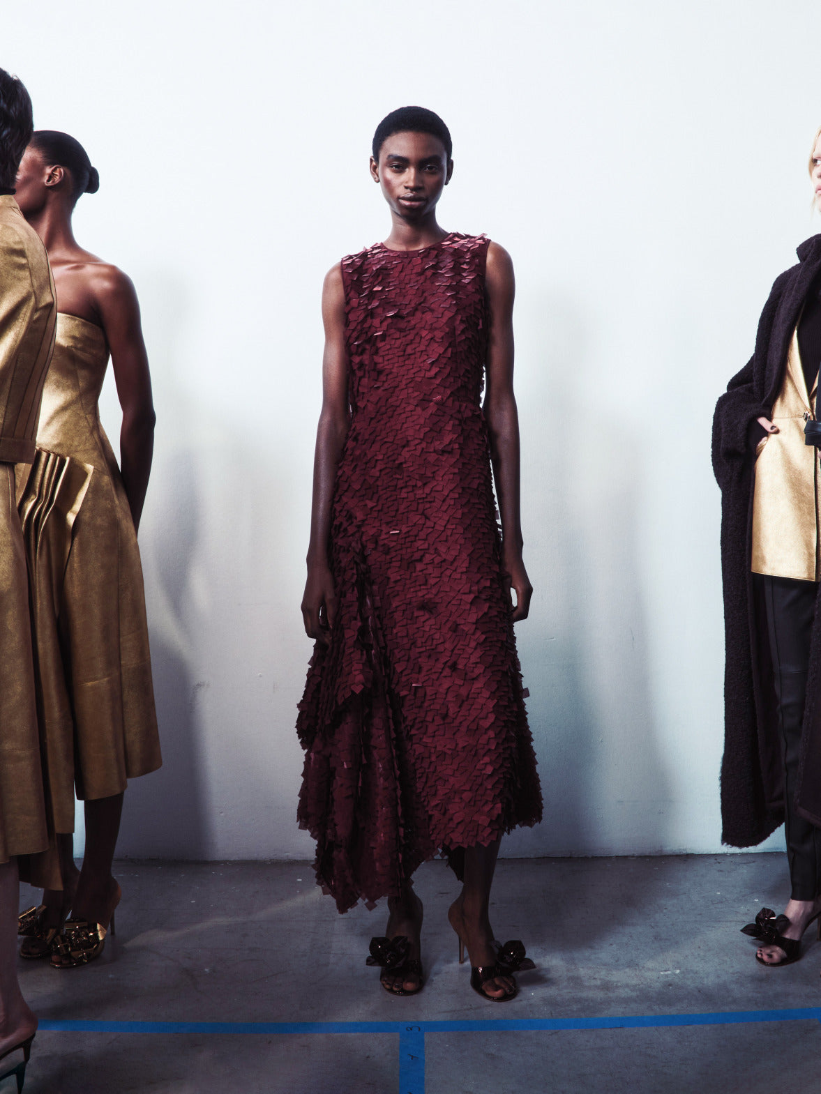 Ein Model steht in einem burgunderroten Kleid mit lasergeschnittener 3D-Textur, flankiert von Models in metallisch-goldenen und schwarzen strukturierten Gewändern.
