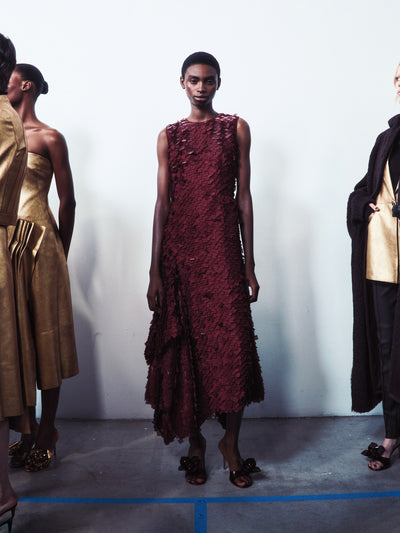 Ein Model steht in einem burgunderroten Kleid mit lasergeschnittener 3D-Textur, flankiert von Models in metallisch-goldenen und schwarzen strukturierten Gewändern.