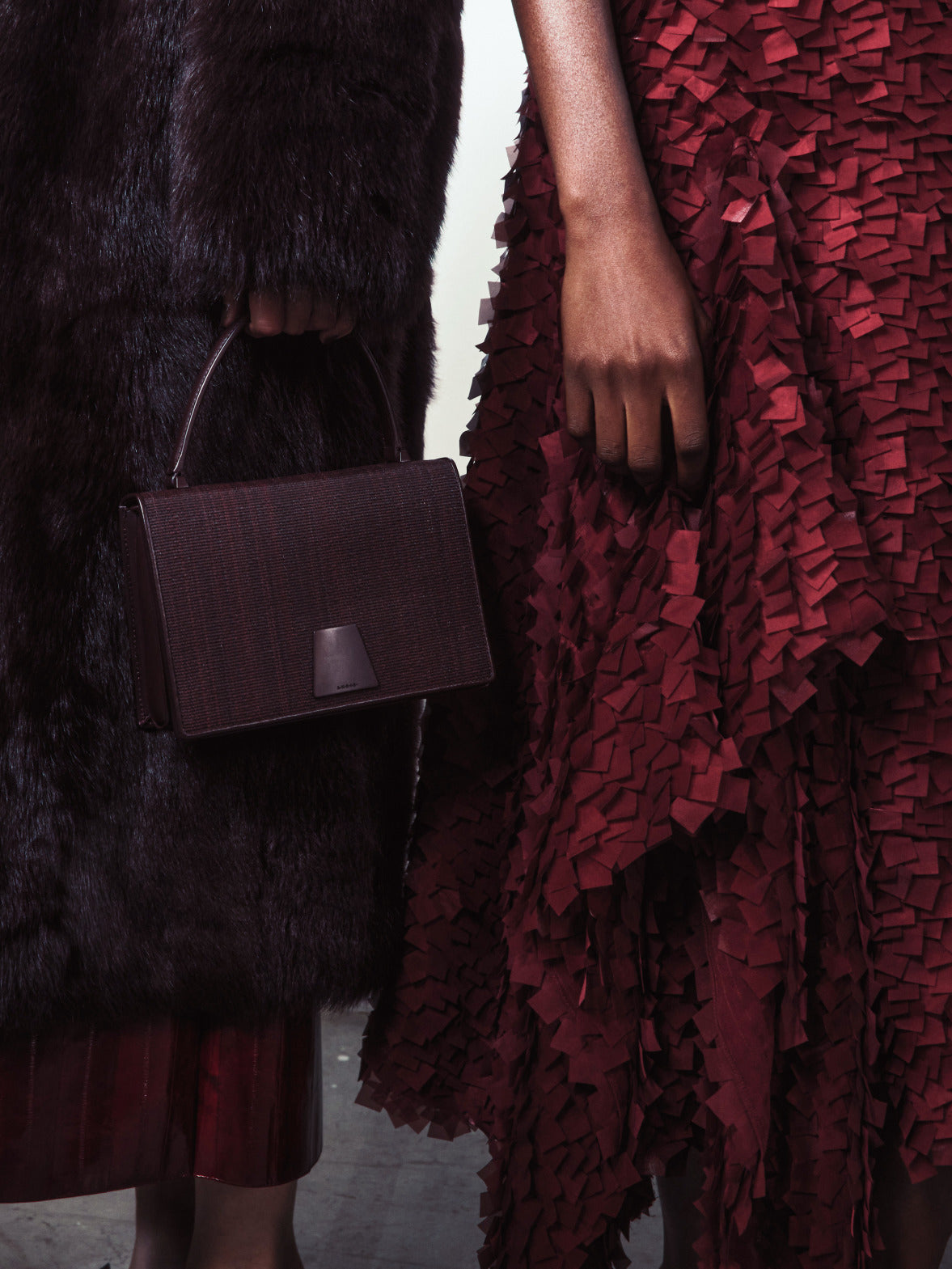 Eine Nahaufnahme einer strukturierten burgunderroten Handtasche vor einem dunklen Pelzmantel und einem Rock mit dichter, lasergeschnittener 3D-Blütenblatt-Textur.