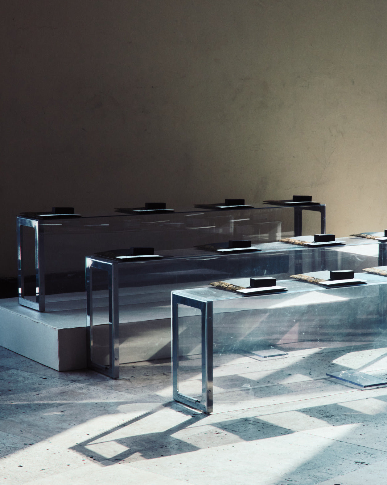 Eine minimalistische Ansicht von transparenten Plexiglasbänken in einem Studio, die das natürliche Licht am Boden reflektieren und kleine schwarze Geschenkkartons für die Gäste bereithalten.
