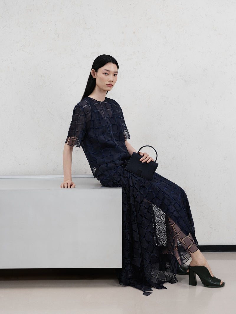 Erleben Sie die Abendgala-Eleganz della Akris Pre-Fall Kollektion mit einer tiefblauen Robe, die durch ein kunstvolles, transparentes Karomuster fasziniert. Perfekt ergänzt durch eine strukturierte, schwarze Luxushandtasche strahlt dieser Look moderne Raffinesse aus. Entdecken Sie erlesene Texturen und meisterhafte Schweizer Schneiderkunst.