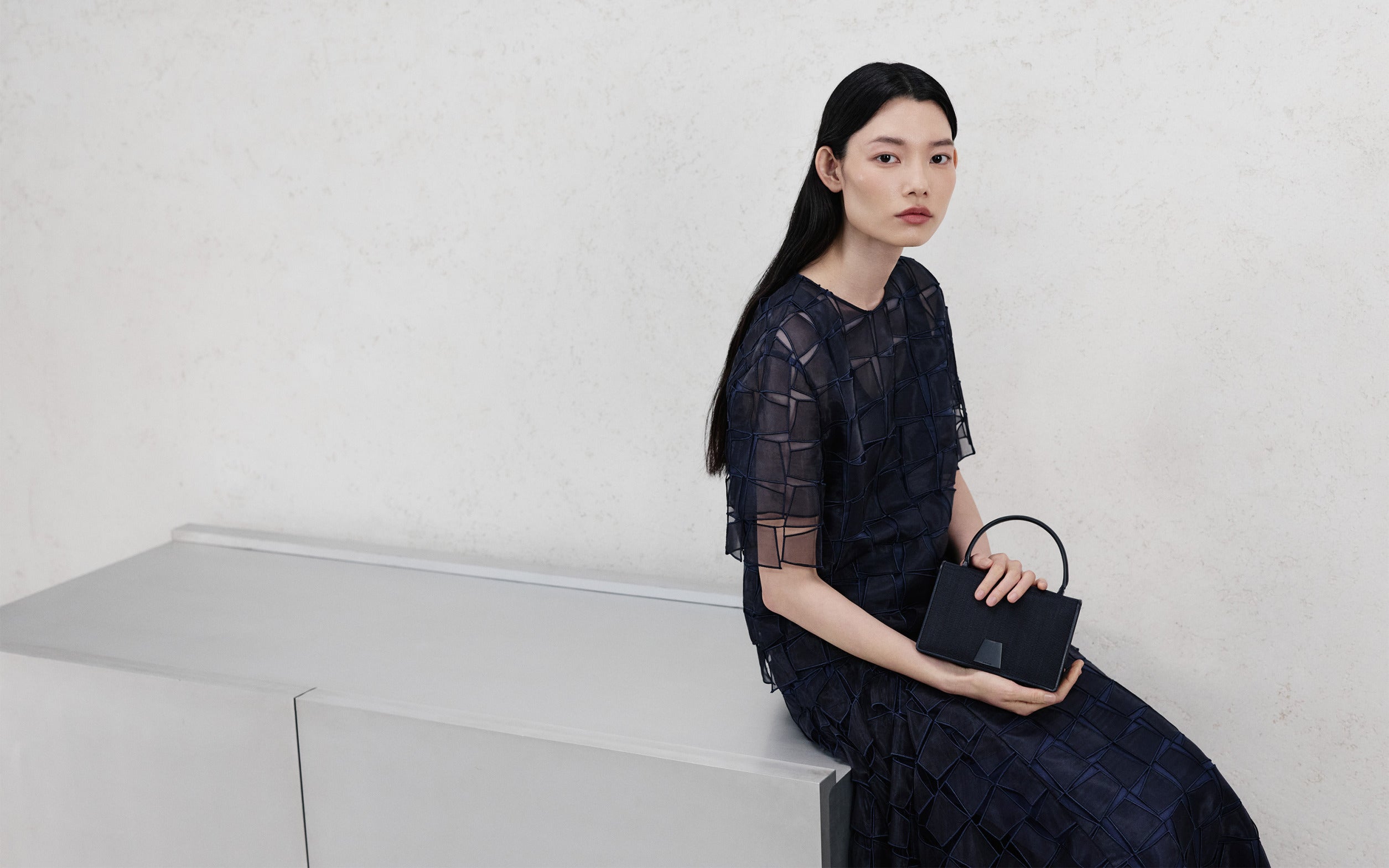 Akris Pre-Fall 2026 Kollektion mit einem transparenten, dunkelblauen Patchwork-Oberteil und passendem Rock. Der Look wird durch eine strukturierte schwarze Lederhandtasche mit Henkel vor einem minimalistischen, cremefarbenen Hintergrund ergänzt.