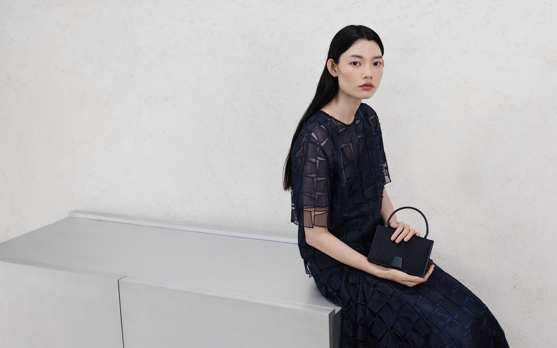 Model in einem dunkelblauen Akris Pre-Fall Kleid mit geometrischer, architektonischer Stickerei und einer passenden Denim-Handtasche, sitzend vor einer minimalistischen Wand.