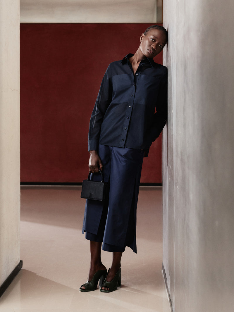 Erleben Sie den raffinierten Minimalismus der Akris Pre-Fall Kollektion mit einem eleganten, monochromen Look in tiefem Marineblau. Das stilvolle Ensemble aus Hemdbluse und weiter Hose wird durch eine strukturierte, schwarze Luxushandtasche perfekt ergänzt. Entdecken Sie fließende Schneiderkunst und zeitlose Schweizer Eleganz.