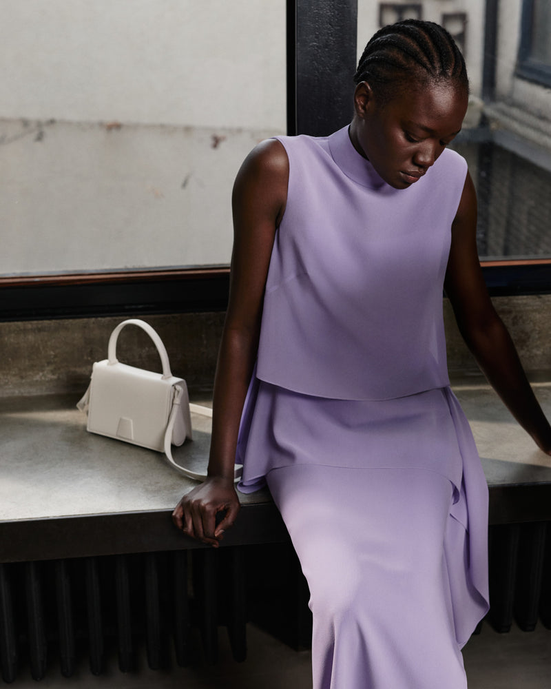 Akris Pre-Fall 2026 Luxusmode-Kollektion präsentiert ein lavendelfarbenes, ärmelloses Top con Stehkragen.
Der dazu passende, drapierte lavendelfarbene Rock kreiert ein elegantes monochromes Ensemble.
Ergänzt durch eine strukturierte cremeweiße Lederhandtasche mit Henkel und Schulterriemen.