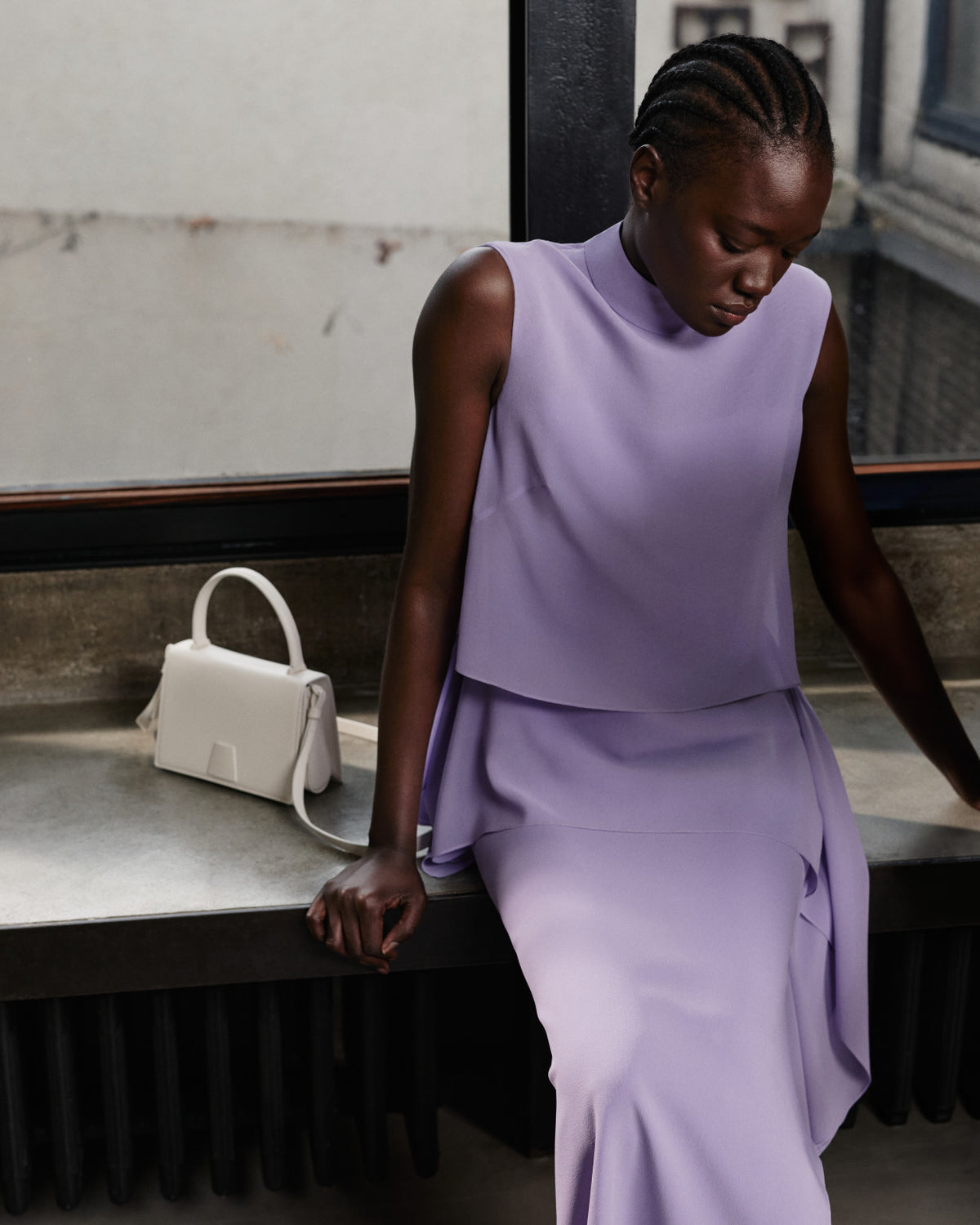 Model in einem malvenfarbenen Akris Pre-Fall Kleid, sitzend an einem Fenster neben einer strukturierten weißen Schultertasche aus Leder.