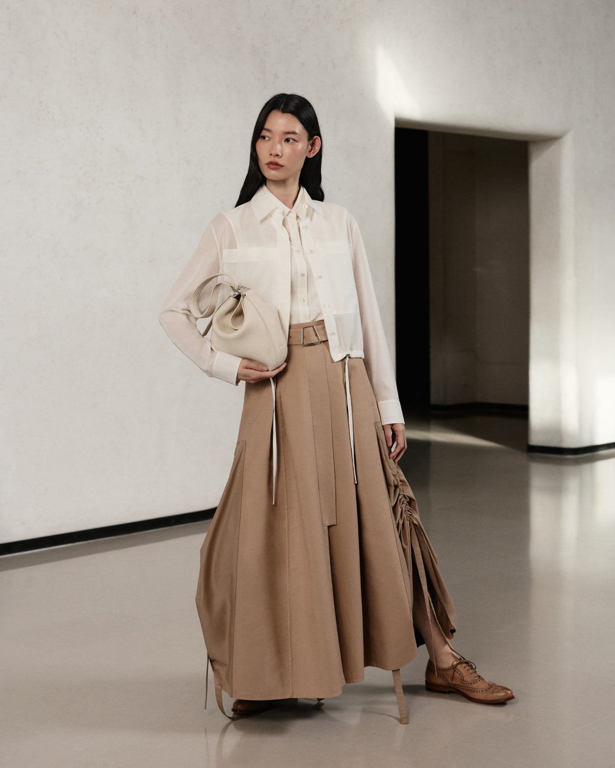 Luxus-Outfit aus der Akris Pre-Fall 2026 Kollektion für Damen.
Weiße, transparente Langarmbluse kombiniert mit einem beigen Maxi-Rock mit Gürtel.
Ergänzt durch eine passende cremefarbene Lederhandtasche und klassische braune Brogues