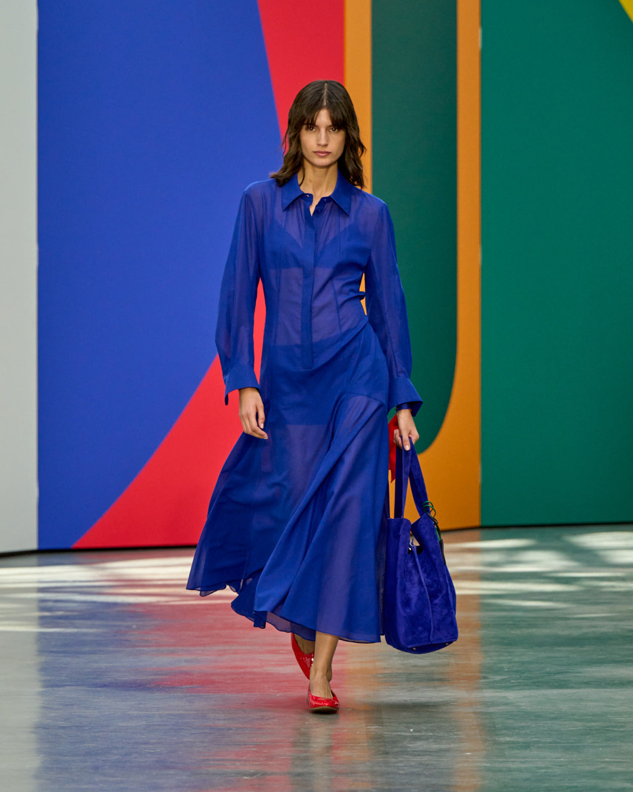 Ein Model läuft den Runway in einem transparenten kobaltblauen Hemdkleid mit langen Ärmeln, trägt eine passende blaue Wildledertasche und rote Flats, vor einem kräftigen mehrfarbigen geometrischen Hintergrund.