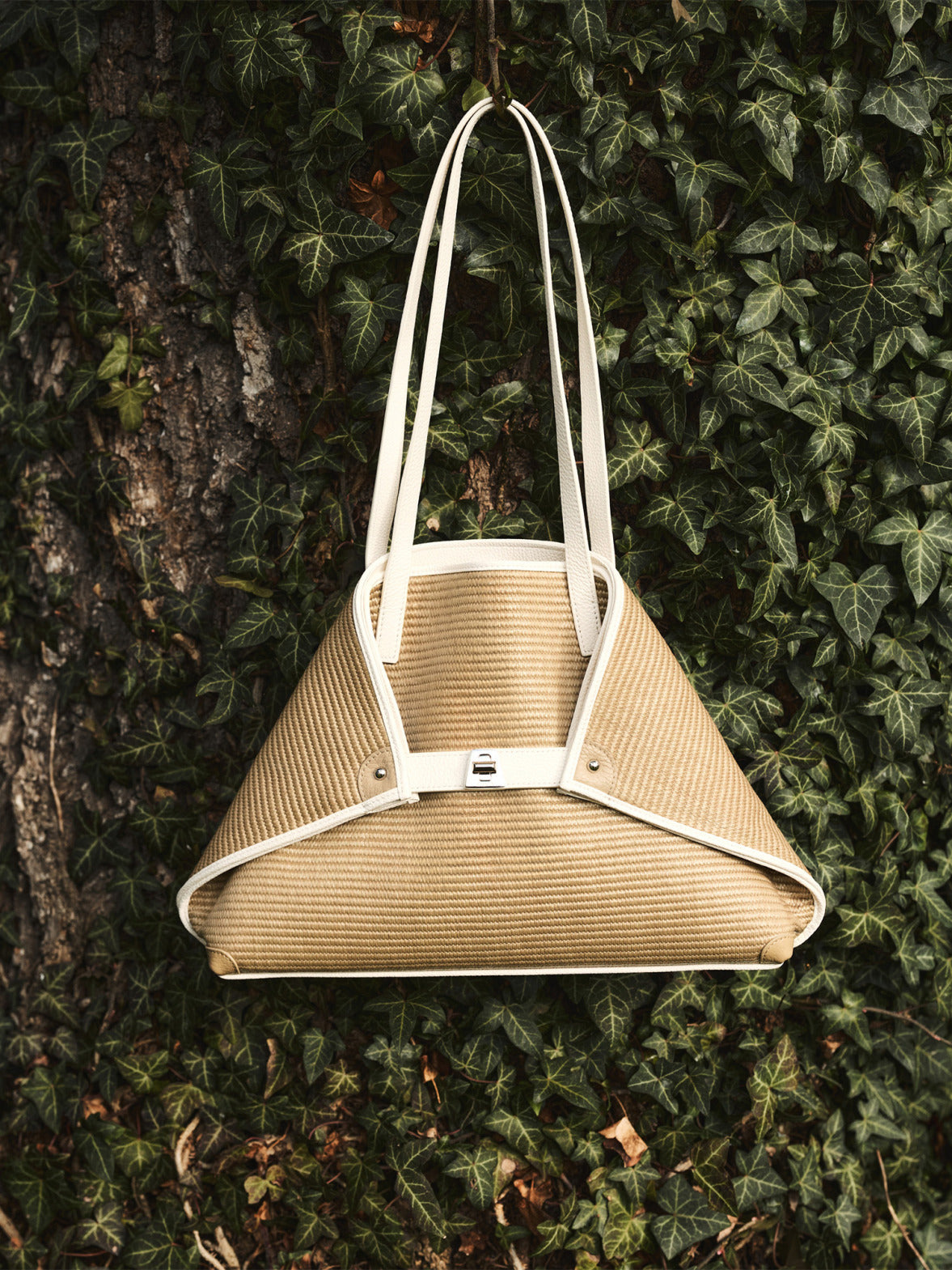 Akris Handtasche aus geflochtenem Bast mit kontrastierenden weißen Lederpaspeln und Henkeln.

Dreieckige Designer-Tragetasche von Akris, aufgehängt an einem efeu-bewachsenen Baum.