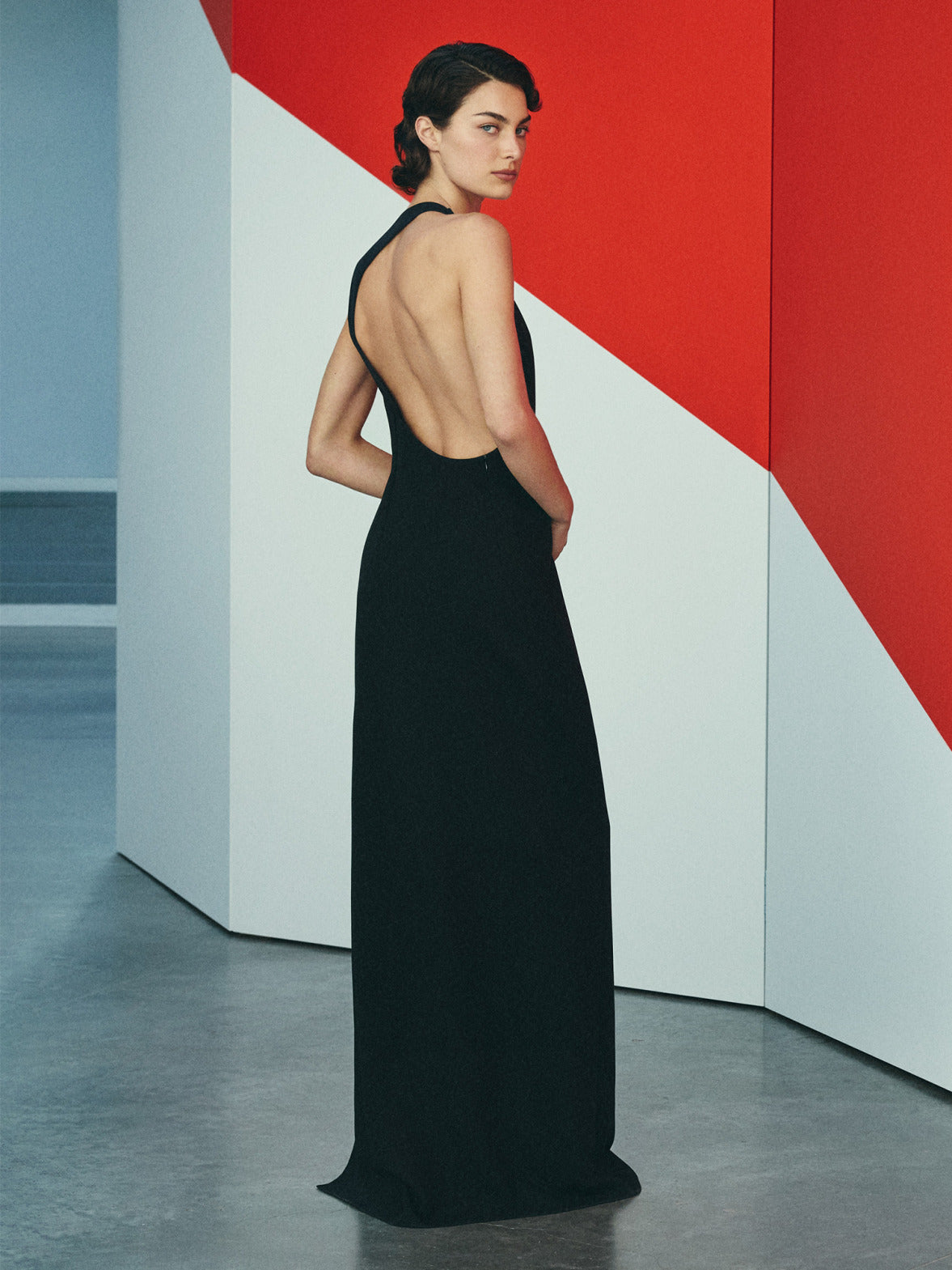 Ein Model in einem eleganten, rückenfreien schwarzen Kleid vor einem geometrischen, rot-weißen Hintergrund.
Die Aufnahme betont die minimalistische Ästhetik und die klare Schnittführung des Kleides.