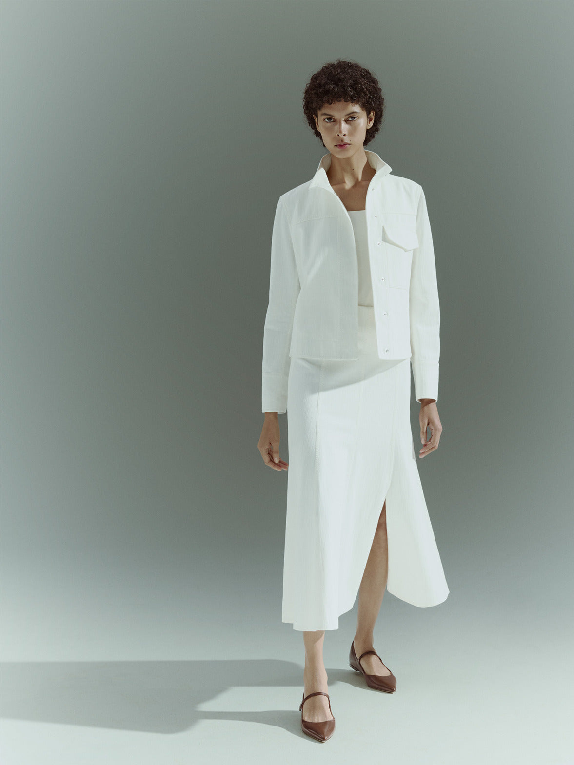 Modernistischer Akris punto Pre-Fall 2026 Editorial-Look: Eine Frau in einem komplett weißen Textil-Outfit mit Faltenrock sitzt im Profil. Sie trägt minimalistische schwarze, spitze Mary-Jane-Flachschuhe aus Leder vor einem neutralen Studiohintergrund.