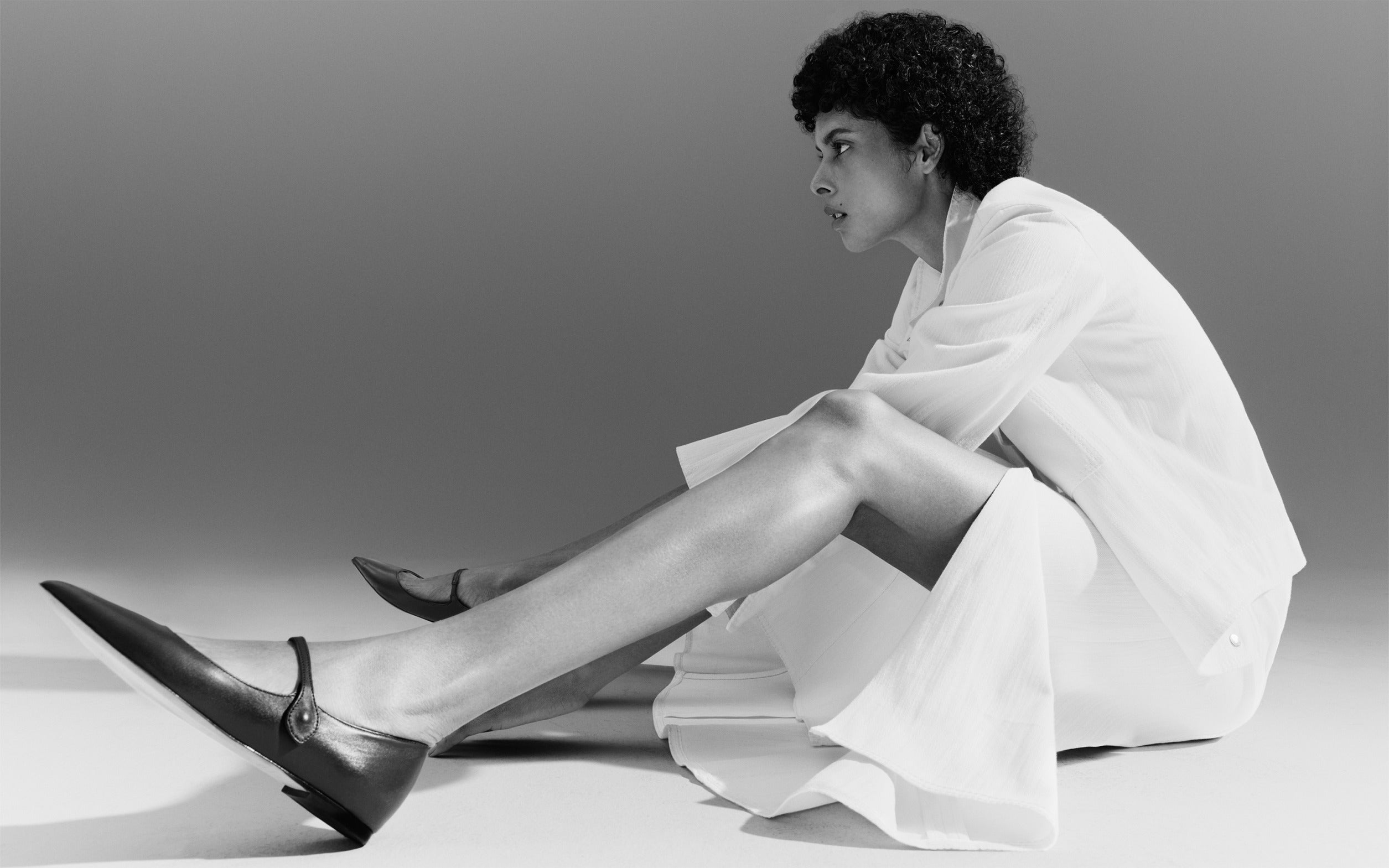 Modernistischer Akris punto Pre-Fall 2026 Editorial-Look: Eine Frau in einem komplett weißen Textil-Outfit mit Faltenrock sitzt im Profil. Sie trägt minimalistische schwarze, spitze Mary-Jane-Flachschuhe aus Leder vor einem neutralen Studiohintergrund.