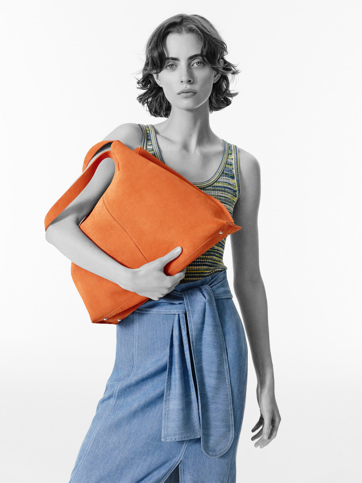 Ein Diptychon eines Models, das zwei farbenfrohe Handtaschen präsentiert: eine große orangefarbene Wildleder-Tote und eine hellblaue geflochtene Umhängetasche mit Fransen.
Die leuchtenden Accessoires kontrastieren mit dem minimalistischen, hellen weißen Hintergrund.