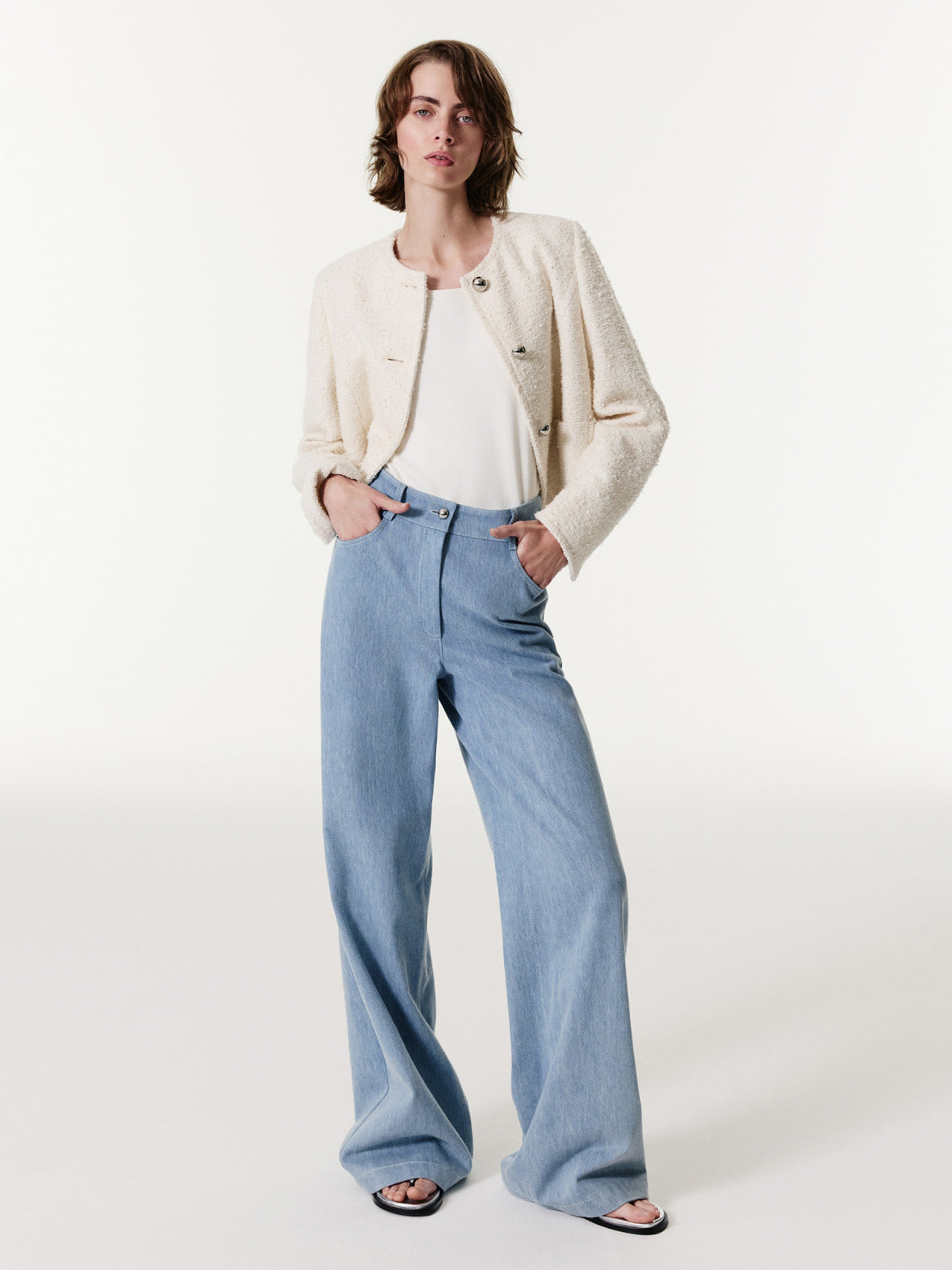 Schickes Model in cremefarbener Bouclé-Strukturjacke und hellblauen Wide-Leg-Jeans, präsentiert ein modernes Ensemble mit Sandalen vor weißem Studiohintergrund.