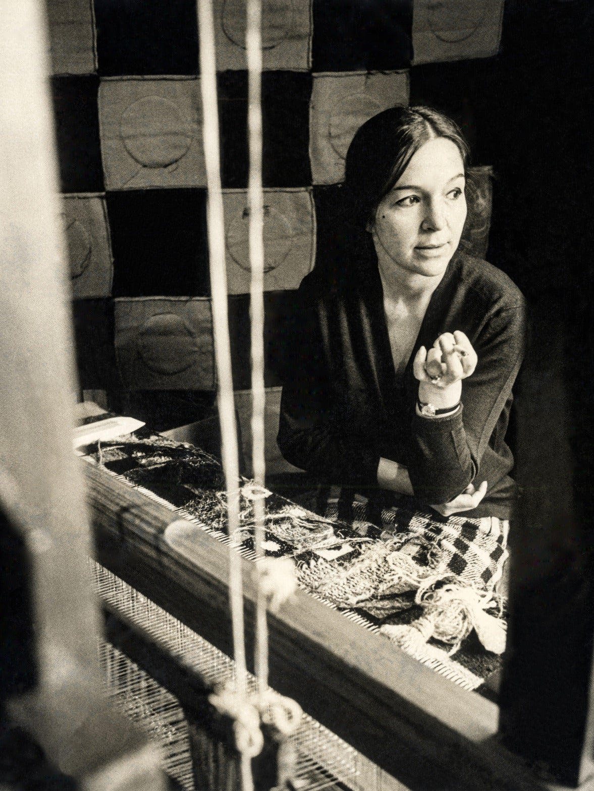 Ein nostalgisches Schwarz-Weiß-Porträt der Künstlerin Olga de Amaral an ihrem Webstuhl, das einen Moment kreativer Reflexion inmitten gewebter Texturen einfängt.